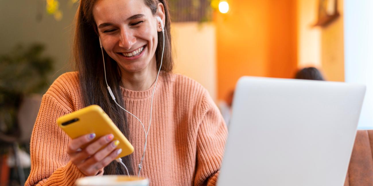 Uma mulher sorrindo em frente a um notebook enquanto segura um celular, feliz por conhecer o programa de cashback