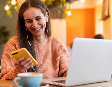 Uma mulher sorrindo em frente a um notebook enquanto segura um celular, feliz por conhecer o programa de cashback